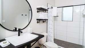 Un baño moderno con un espejo redondo grande, un lavamanos blanco, una encimera negra, un inodoro y una zona de ducha con puerta de cristal.