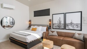 Un dormitorio moderno con una cama, un sofá de cuero marrón, dos cuadros enmarcados de paisajes desérticos, un espejo redondo y un aire acondicionado instalado en la pared.