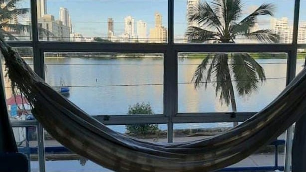 Eine Hängematte, zwischen Fensterrahmen gespannt, mit Blick auf einen Stadt-Horizont und ein Gewässer.