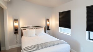 Un dormitorio ordenado con una cama grande, dos lámparas de pared, un ventanal con persianas y un equipo de aire acondicionado.