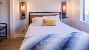 Ein ordentlich bezogenes Bett mit weißer und blauer Decke, zwei Wandlampen und ein Fenster zur Rechten.