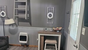 Une pièce avec un plafond en bois, un radiateur, un panier pour animal de compagnie, une table avec une chaise, une porte avec des stores et un petit tapis.