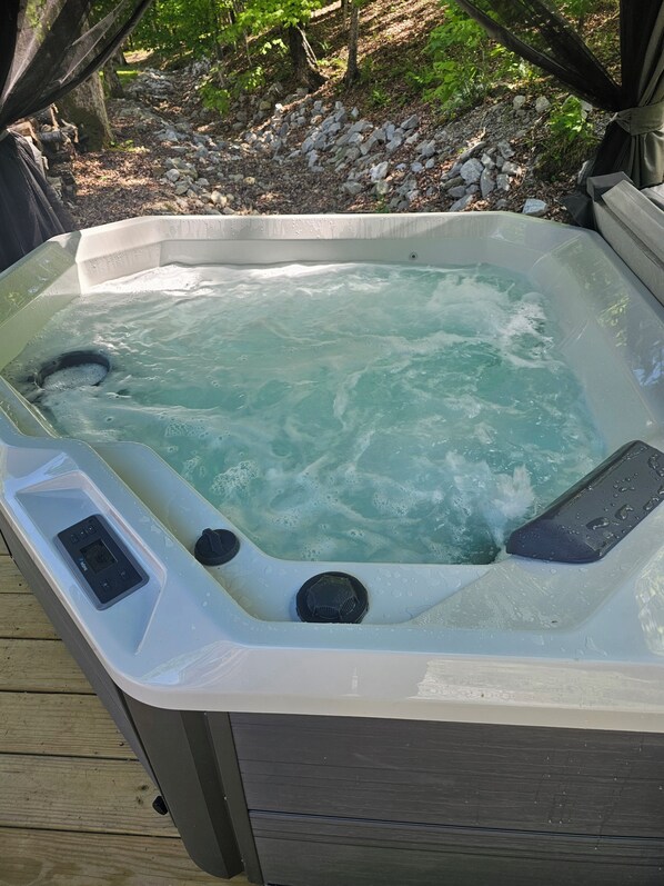 Un jacuzzi con panel de control, rodeado por una terraza de madera y un paisaje natural.