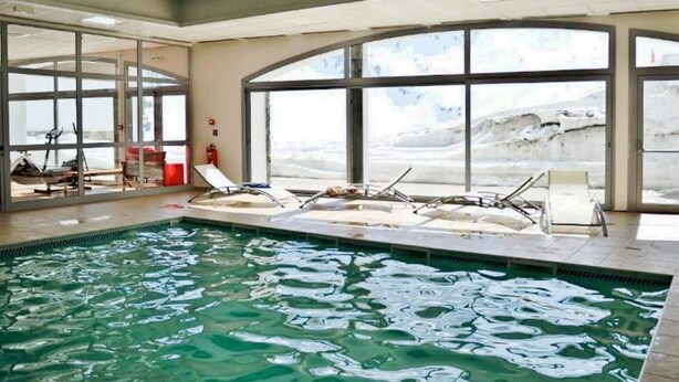Piscina interior com paredes e janelas de vidro transparente, cadeiras de descanso e um extintor de incêndio.