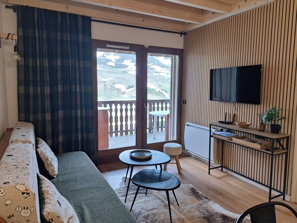 A living room with a sofa, a small table, a TV, and a balcony with a view of the outdoors.