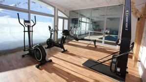Uma academia espaçosa com equipamentos de cardio, uma máquina de remo e amplas janelas com vista para o exterior.