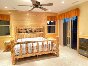 Una cama de madera con tablero de cabecera con motivos forestales, un ventilador de techo y vistas al exterior a través de amplios ventanales.