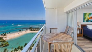 En balkon med udsigt over strand og hav, et rundt træbord, to stole og et billede på væggen.