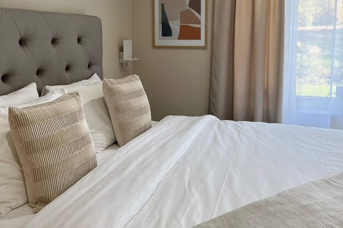 A neatly made bed with a tufted headboard, a framed abstract painting on the wall, and a window with curtains.