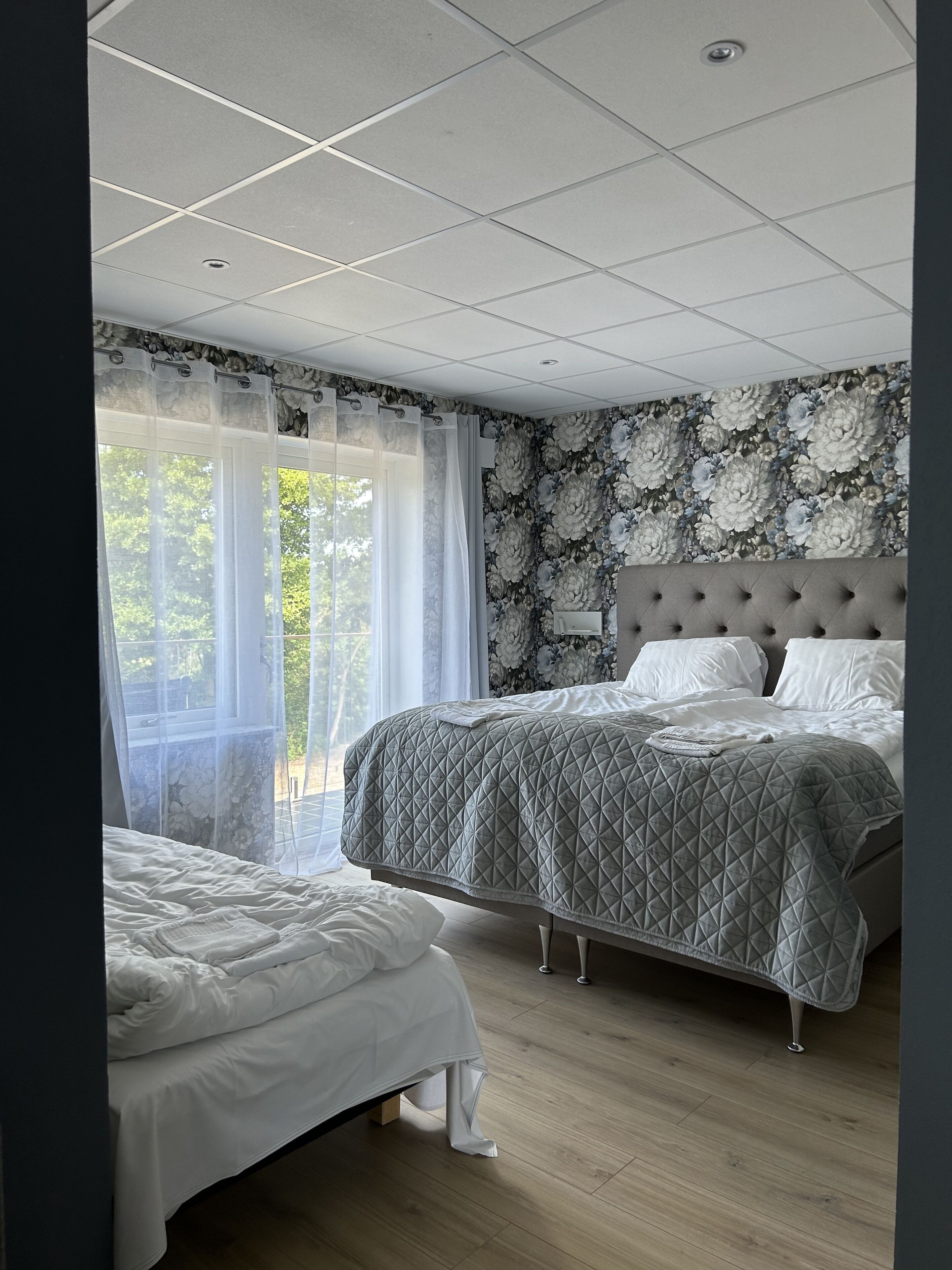 Una camera da letto con due letti, un'ampia finestra con tende leggere, carta da parati a motivi floreali e pavimento in legno.