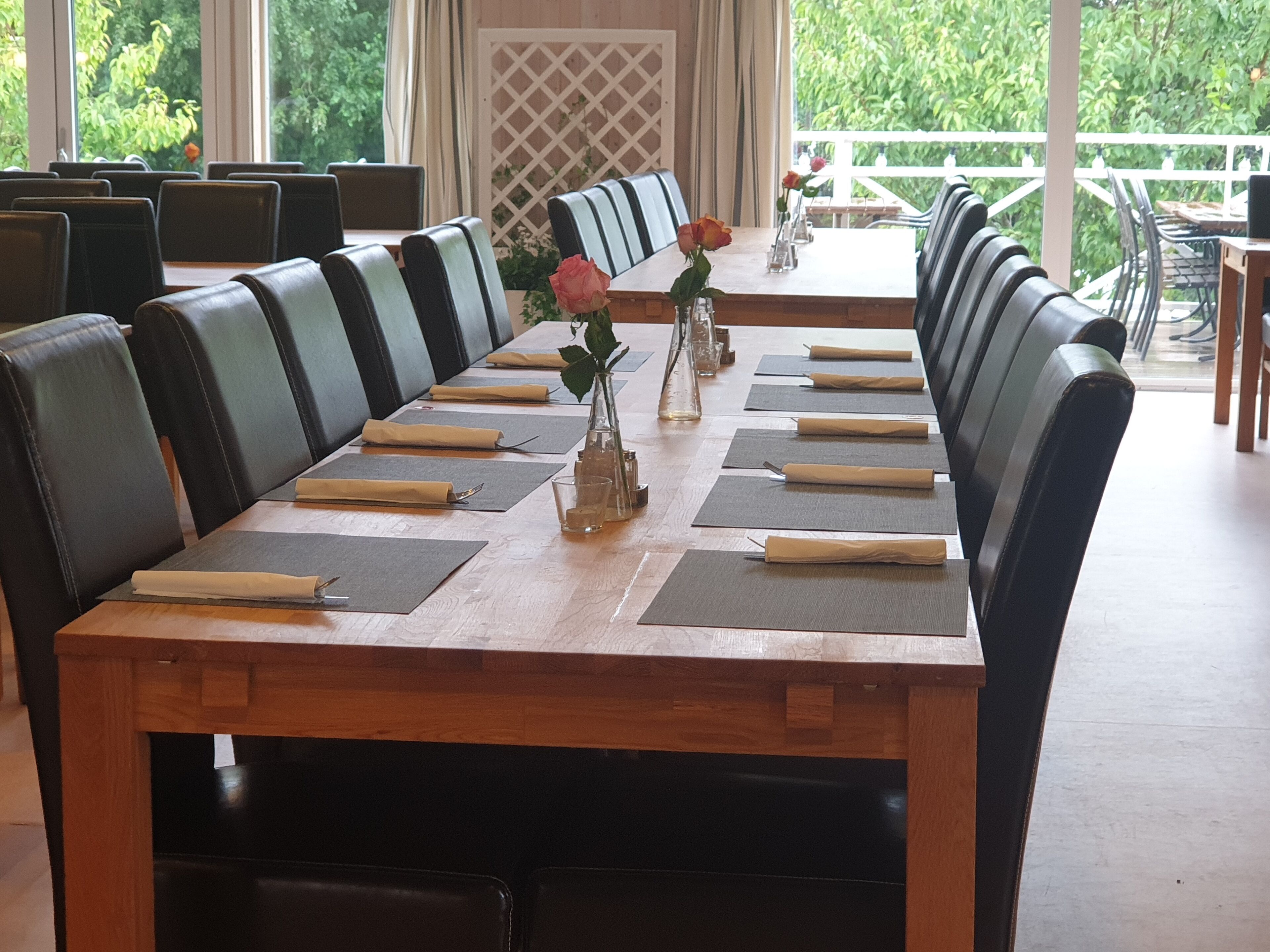 Une longue table à manger, entourée de chaises en cuir noir, est dressée avec soin : chaque place est mise et de petits vases décorés de fleurs y sont disposés.