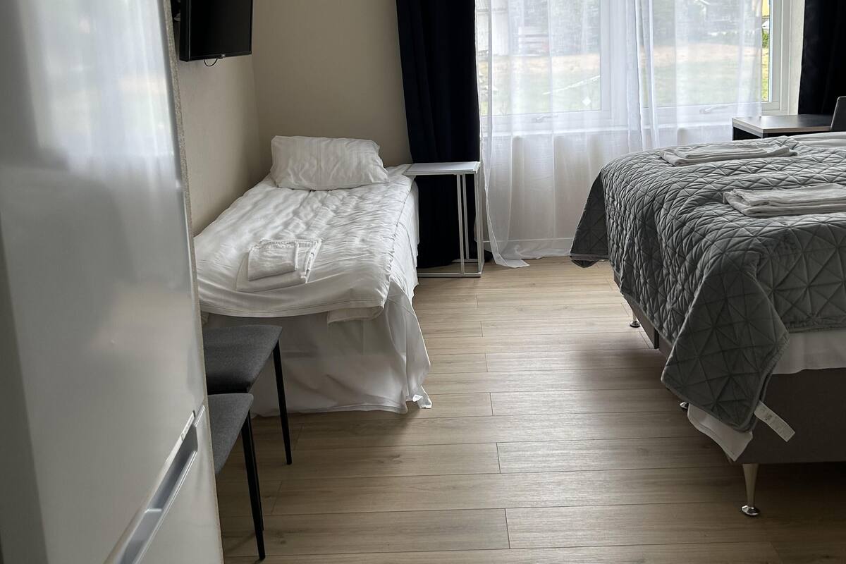 A hotel room with a bed, a desk, a chair, a flat-screen TV, a window with curtains, and a refrigerator.