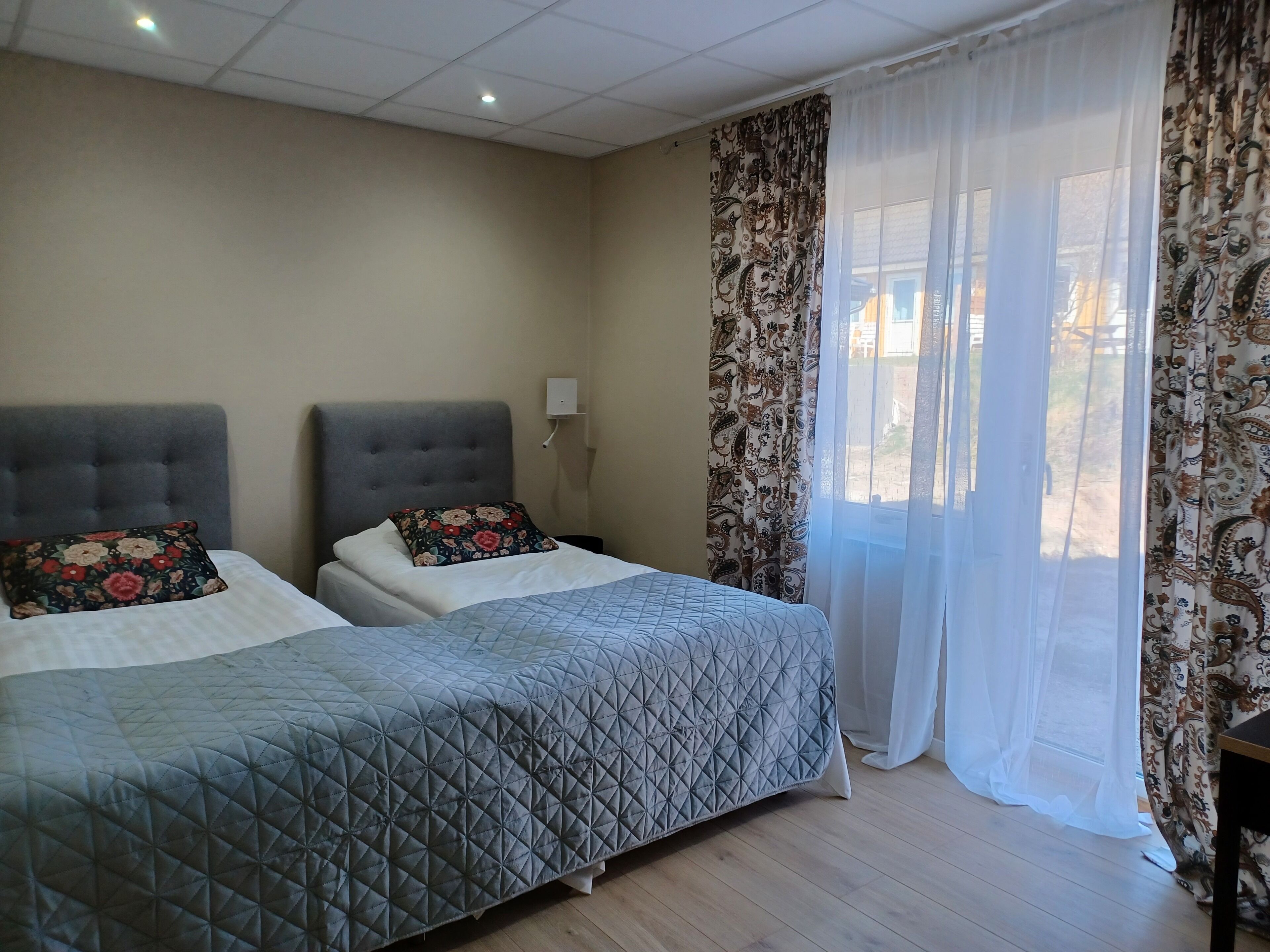 A hotel room with two beds, a large window with sheer curtains, and a light-colored wall.