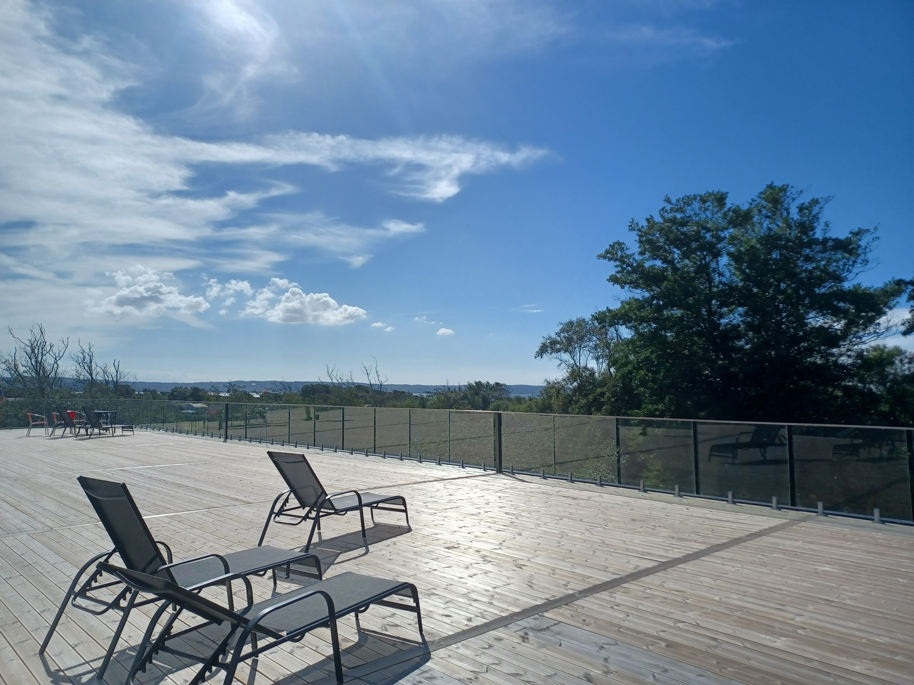 Una terraza de madera con sillones de descanso y barandilla de vidrio, con vista a un paisaje de árboles y colinas al lejano.