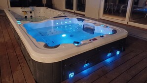 A hot tub with multiple jets and LED lighting, surrounded by a wooden deck and glass doors leading to a patio area. - Hotell Solstickan Mellbystrand (Mellbystrand)