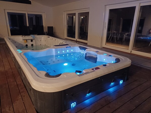 A hot tub with multiple jets and LED lighting, surrounded by a wooden deck and glass doors leading to a patio area.