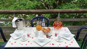 A table set outdoors with a wooden bench, a metal bucket, wine glasses, a water bottle, a basket of croissants, and a potted plant.