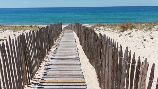 A wooden boardwalk leading to the beach, flanked by sand dunes and a wooden fence.