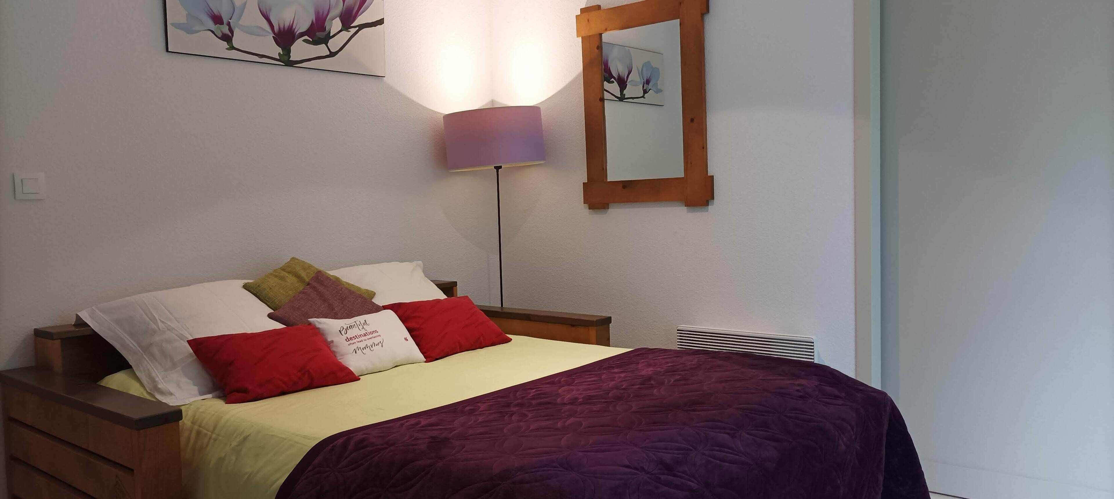 Un lit bien fait, recouvert d’une couverture violette, avec une lampe de chevet et un miroir fixé au mur.