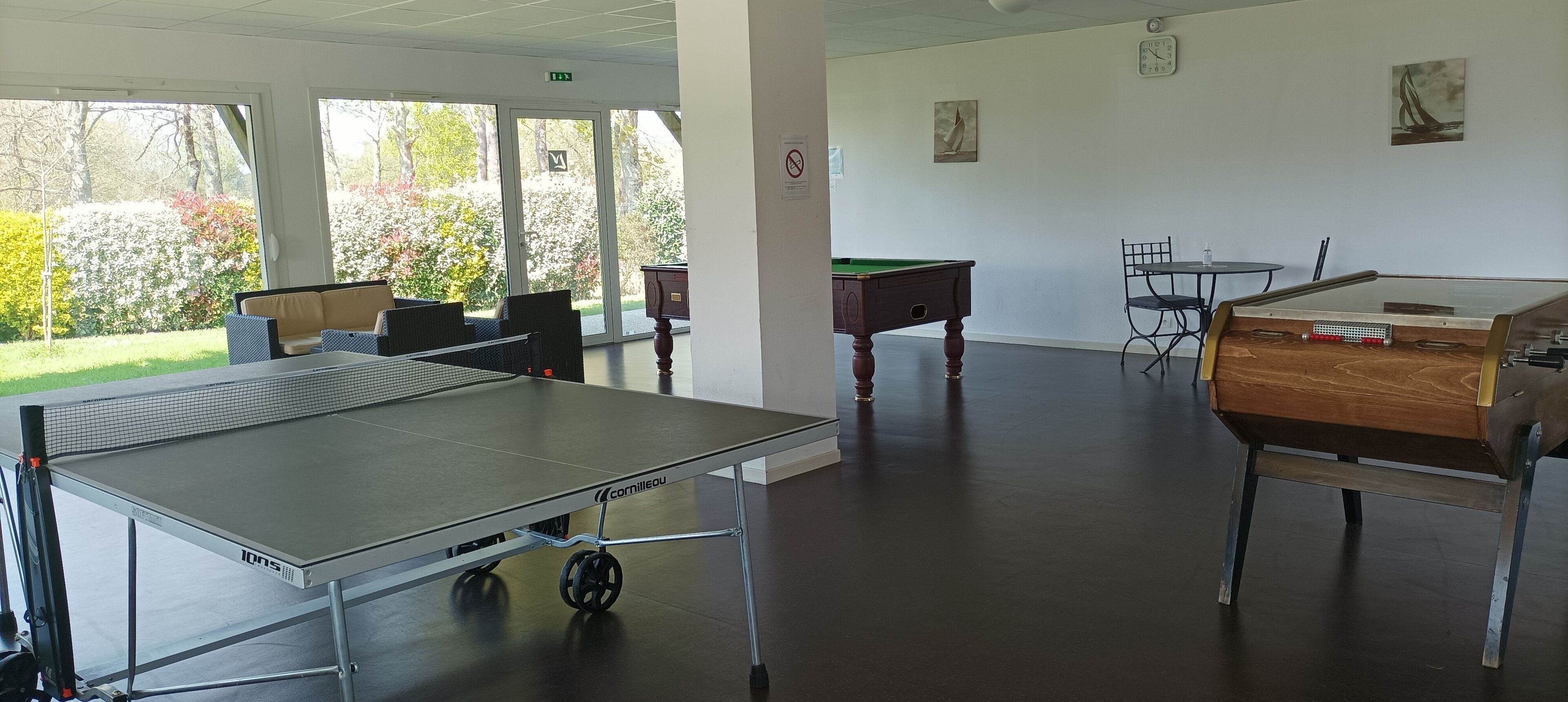 Une pièce équipée d’une table de ping-pong, d’une table de billard et d’une table de jeu de fléchettes. On y trouve des chaises et une table sur laquelle sont posés une horloge et des œuvres d’art accrochées au mur.