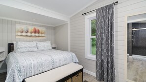 Una camera da letto con un letto, una panca, una finestra con tende e un quadro appeso al muro, oltre a un angolo doccia.
