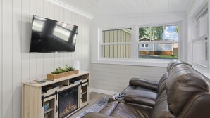 Un soggiorno con TV a schermo piatto montata a parete, mobile TV in legno, divano in pelle e finestra con vista su una casa e alberi.