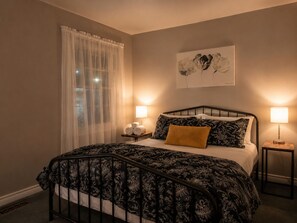 A bedroom with a bed, two bedside lamps, a window with sheer curtains, and a wall-mounted artwork.
