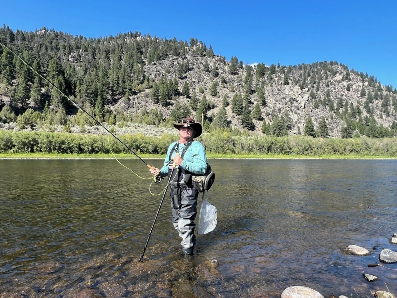 A person standing in a river with a fishing rod, surrounded by a mountainous landscape.
