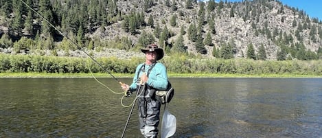 A person standing in a river with a fishing rod, surrounded by a mountainous landscape.