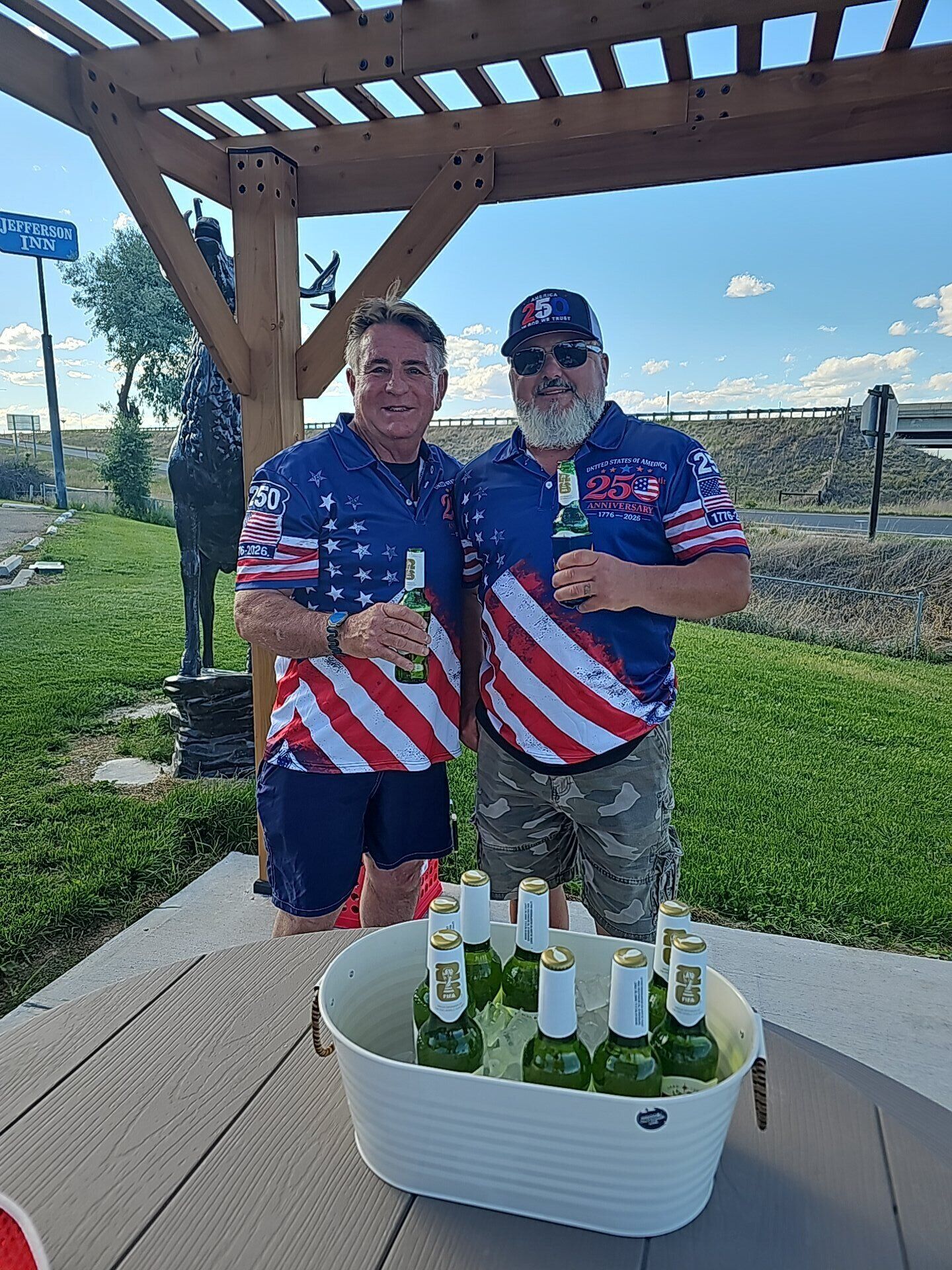 Two people standing under a wooden pergola holding bottles, with a bucket of green bottles in front of them.