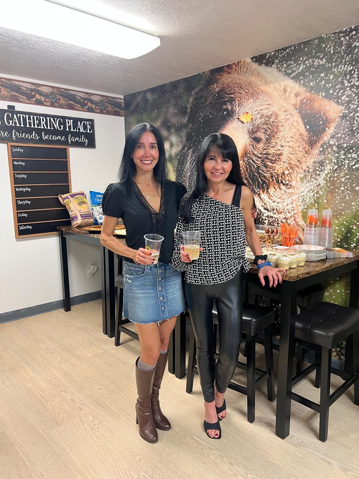 Dos personas de pie junto a una mesa con comida y bebidas, una pared con el letrero 'GATHERING PLACE' y una pizarra de menú que muestra los días de la semana.