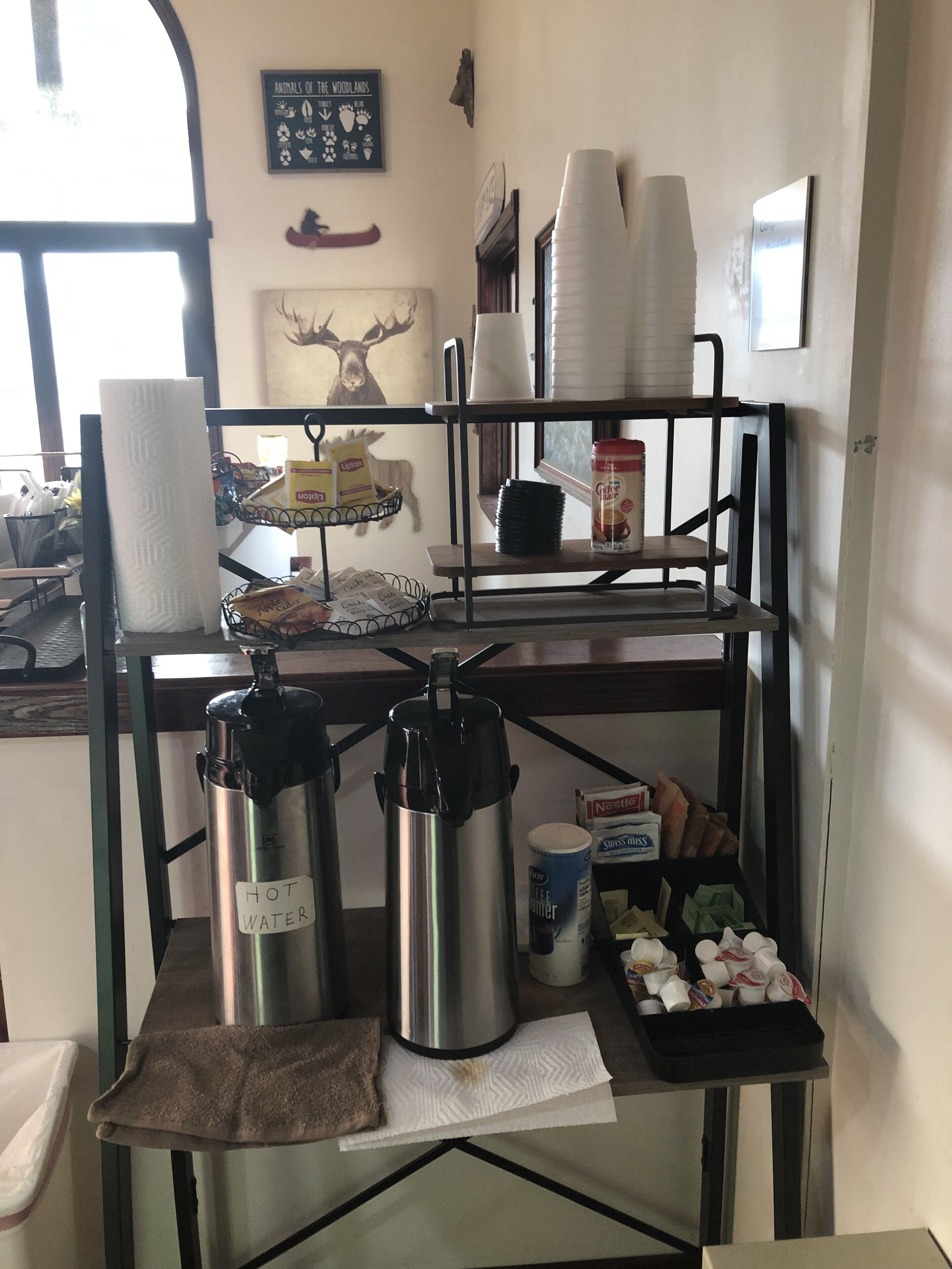 A kitchenette with two stainless steel hot water dispensers, a coffee maker, a stack of disposable cups, and a tray with food items.