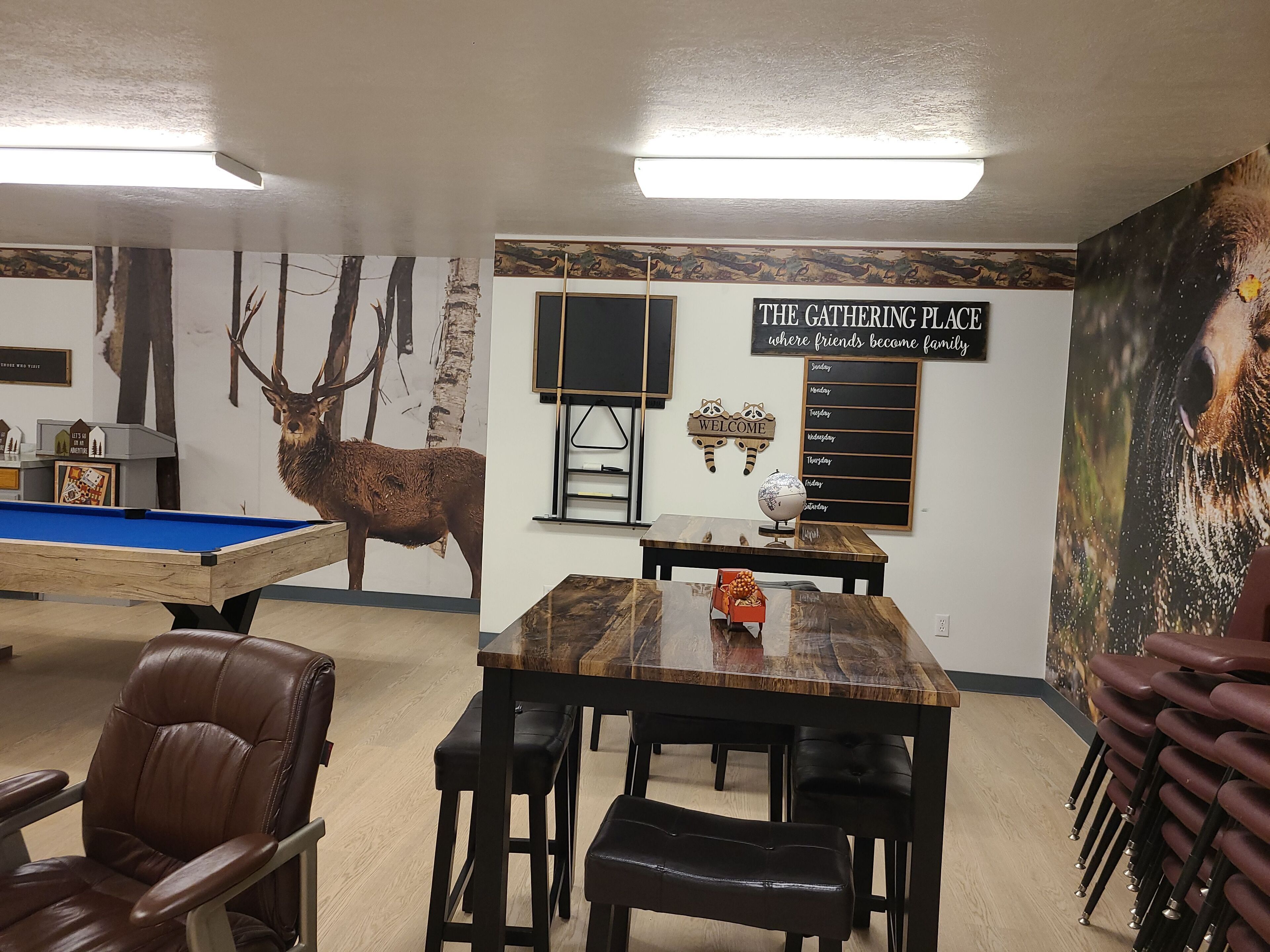Une pièce avec une table de billard, une table à manger en bois avec des bancs, une enseigne indiquant «The Gathering Place» et une fresque murale représentant un cerf.