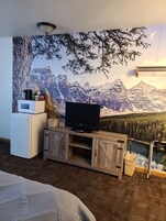 Un dormitorio con un mural de montañas, un televisor sobre un soporte de madera, un mini refrigerador y una cama.