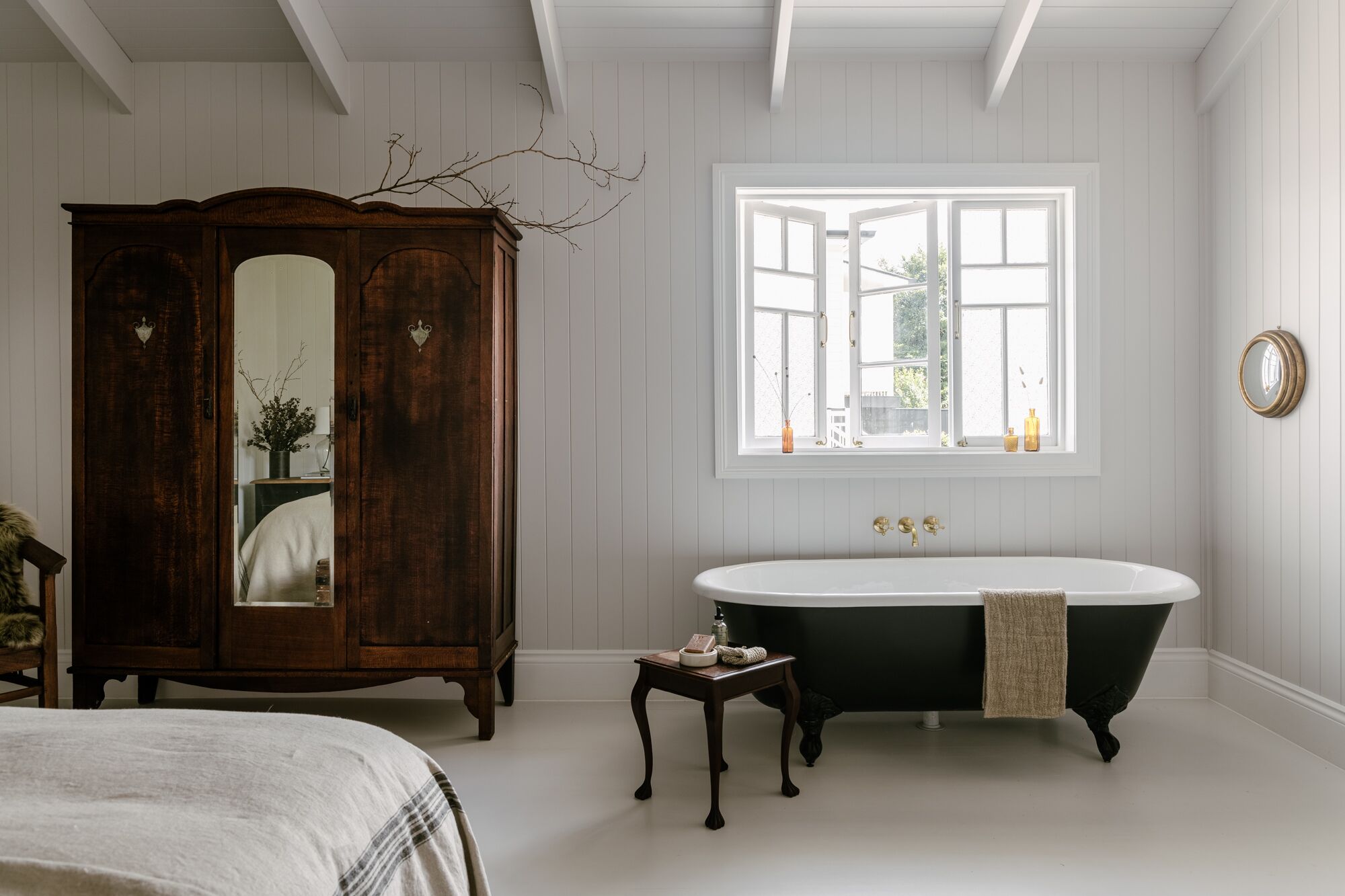Une salle de bain classique avec une baignoire sur pieds à pattes de lion, une armoire en bois dotée d’un miroir et une fenêtre avec des volets blancs.