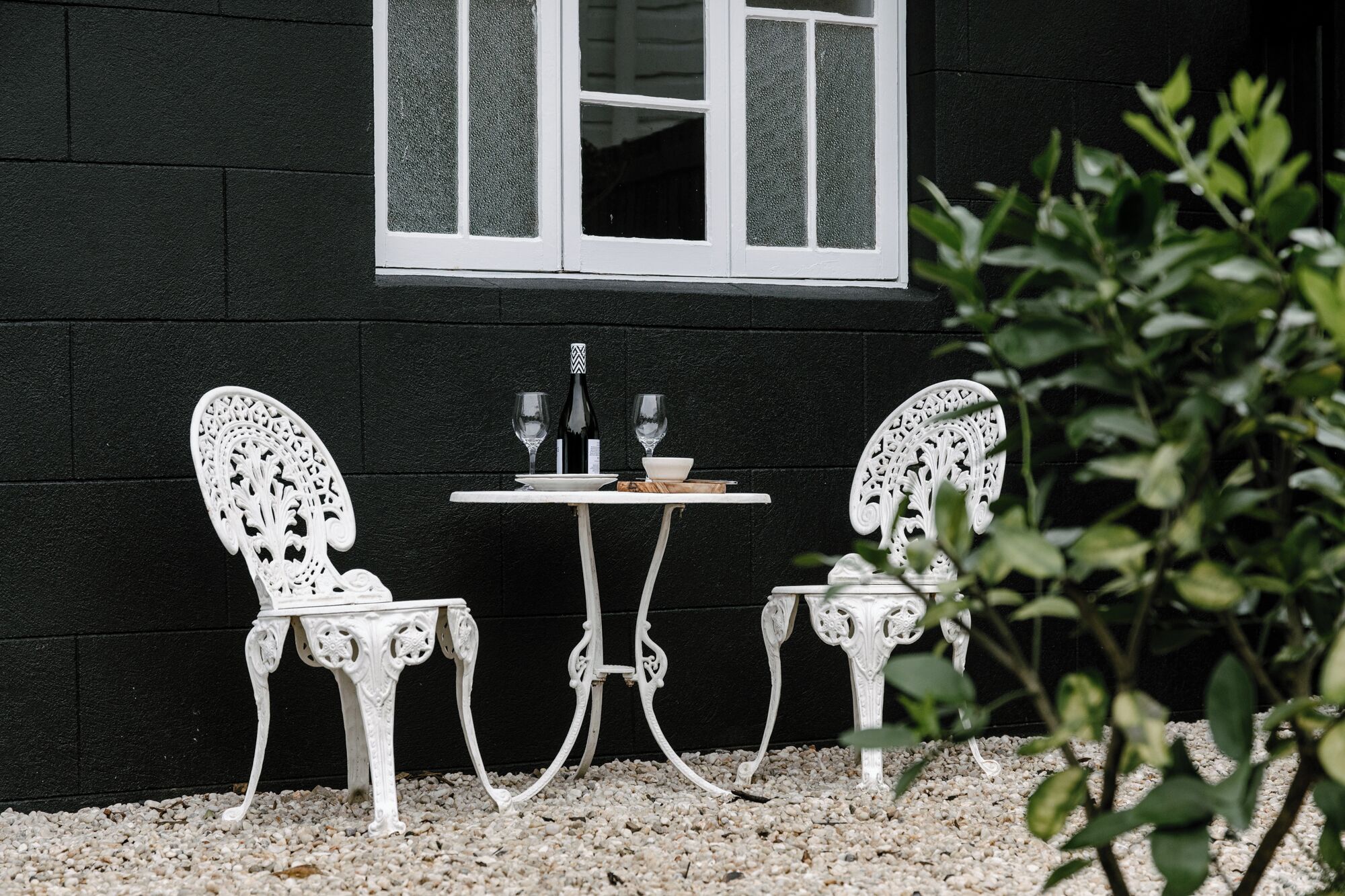 Un espace extérieur aménagé avec des meubles blancs, comprenant deux chaises et une table, placé devant un mur sombre sur lequel se trouve une fenêtre.