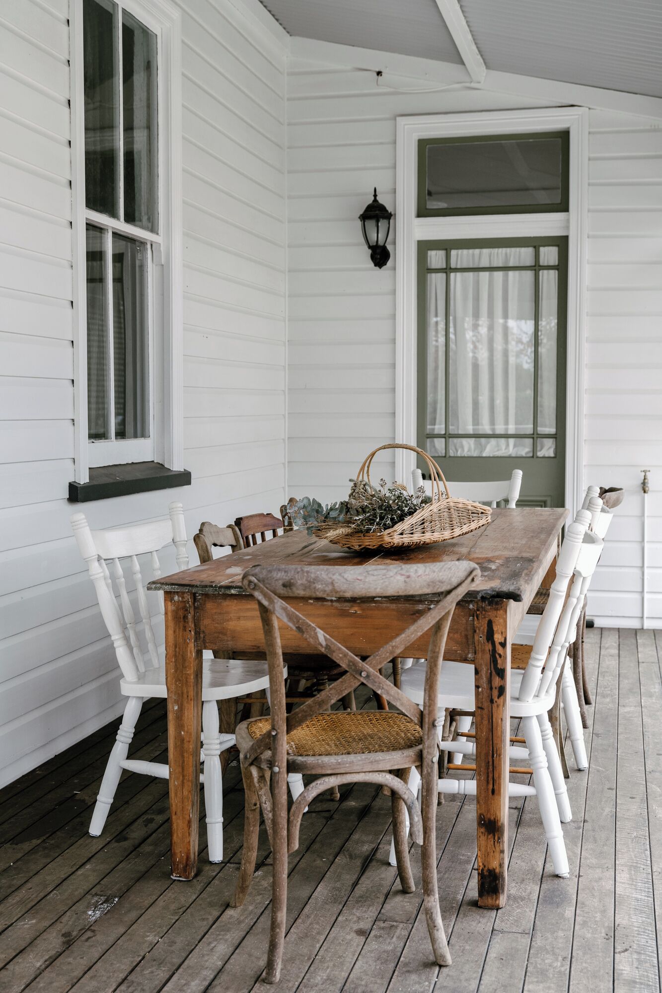 Una mesa de comedor de madera con sillas blancas, una cesta y un farol en una terraza de madera.