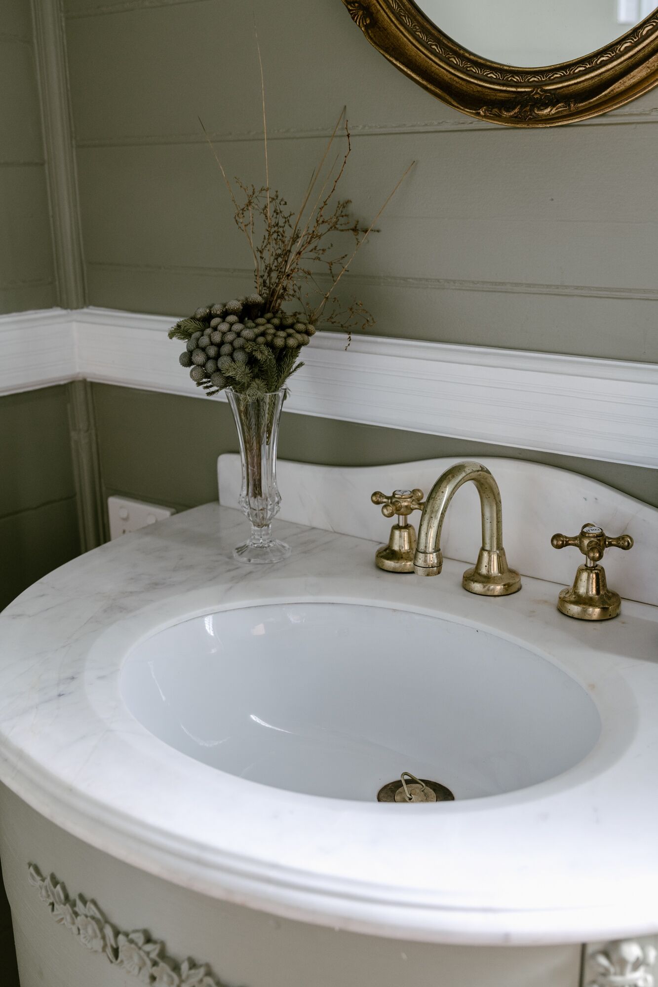 Een badkamer met een wit wastafelmeubel, gouden kranen en een vaas met gedroogde planten.