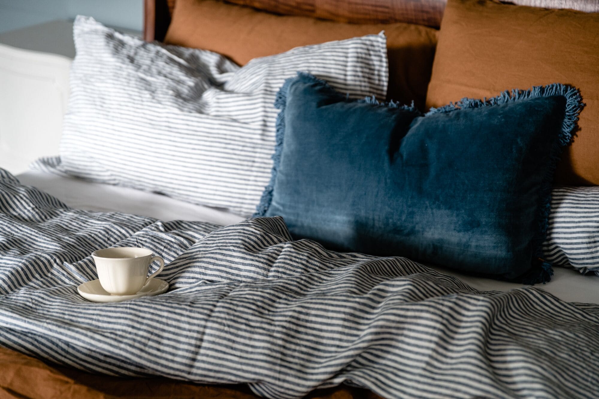A bed with striped bedding, a dark blue pillow, and a white mug on a saucer.