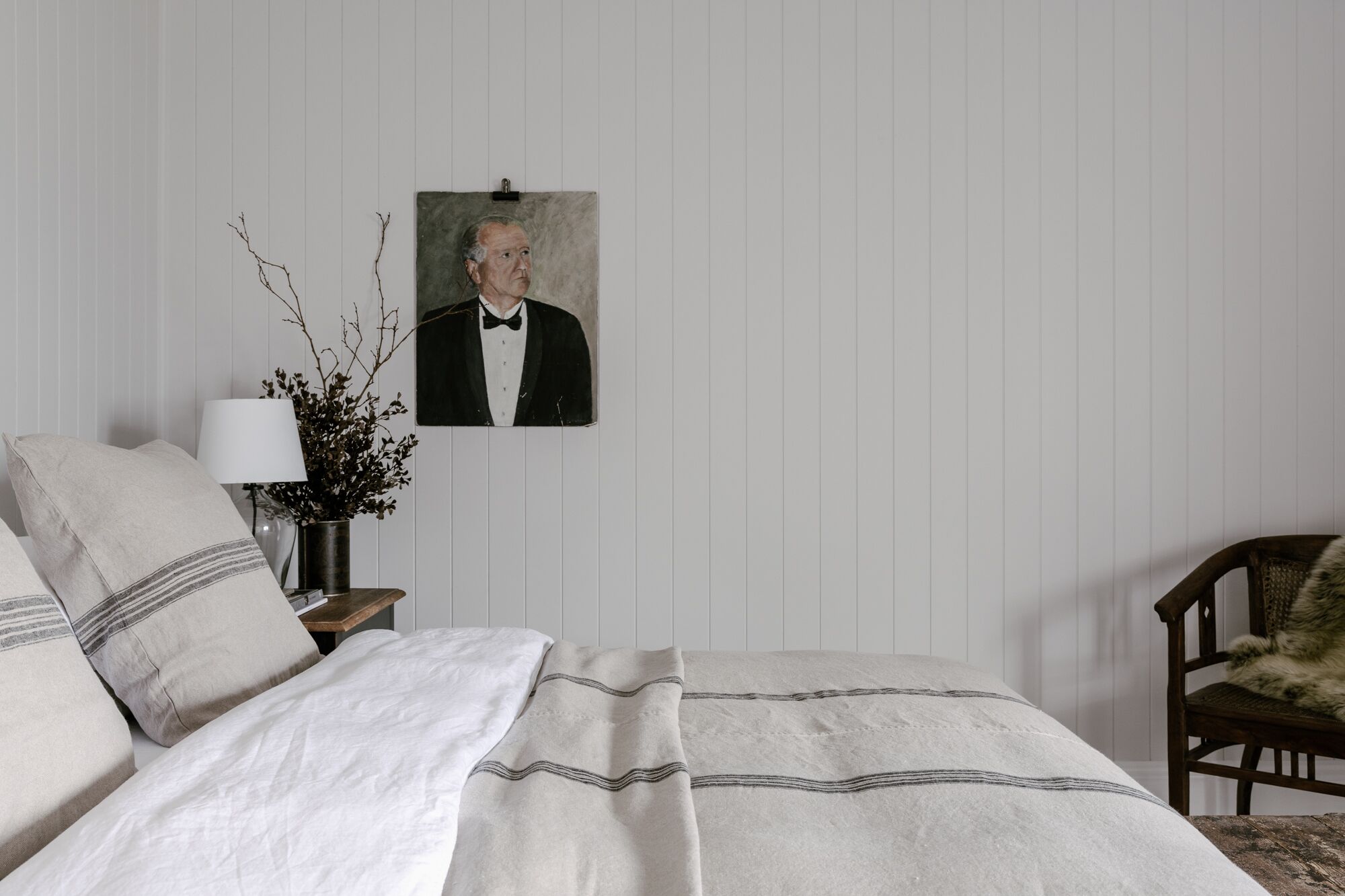 A neatly made bed with a white and gray striped blanket, a framed portrait on the wall, a lamp, and a chair.