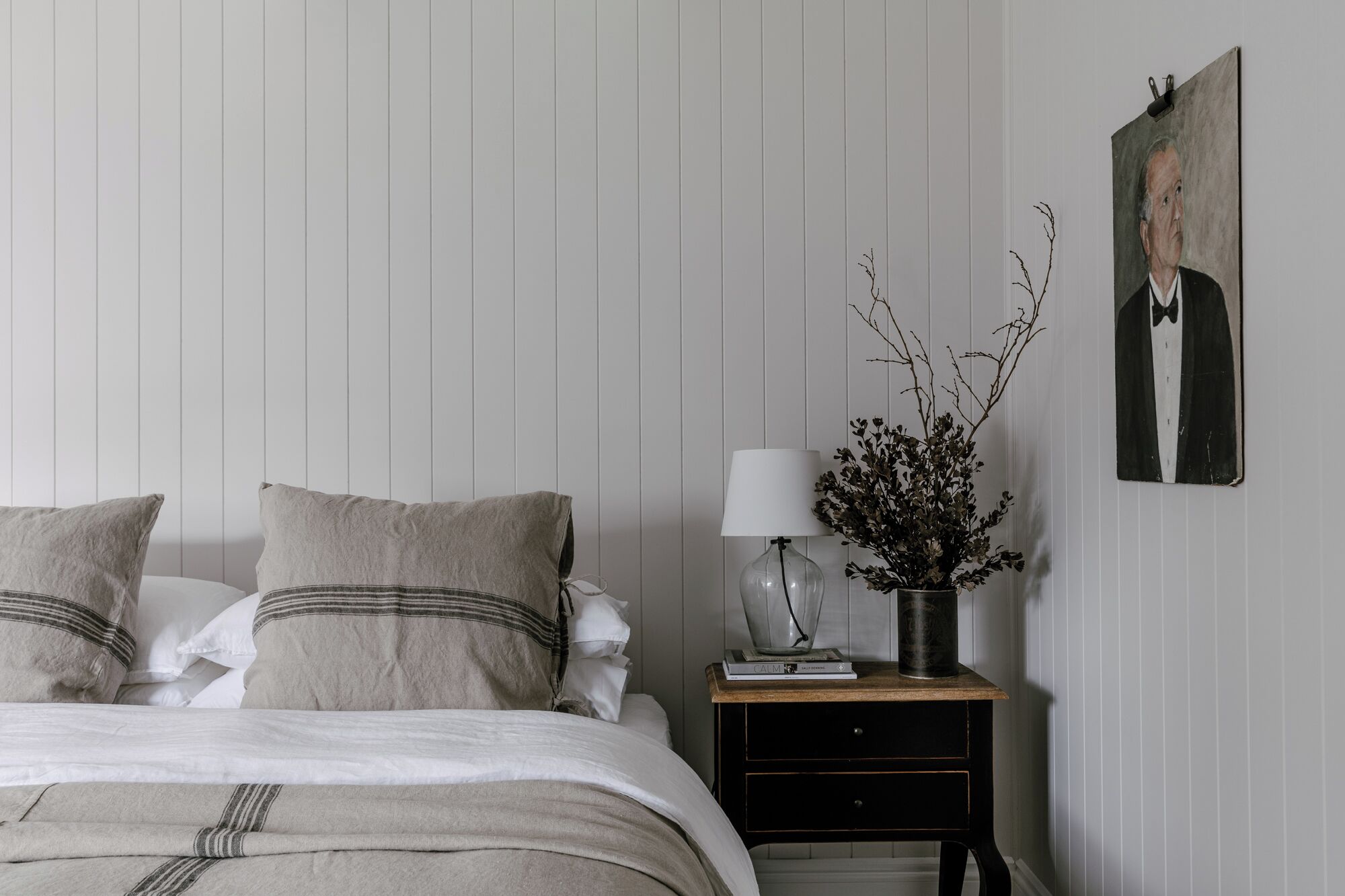 A bedroom with a bed, pillows, a bedside table with a lamp, a vase with branches, and a framed portrait on the wall.