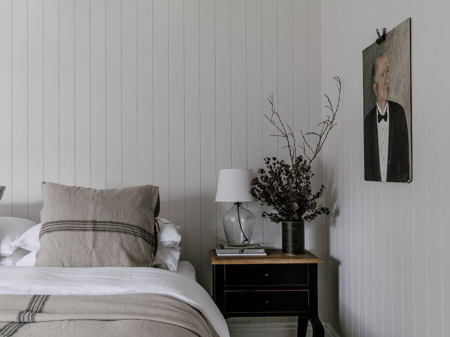 A bedroom with a bed, pillows, a bedside table with a lamp, a vase with branches, and a framed portrait on the wall.