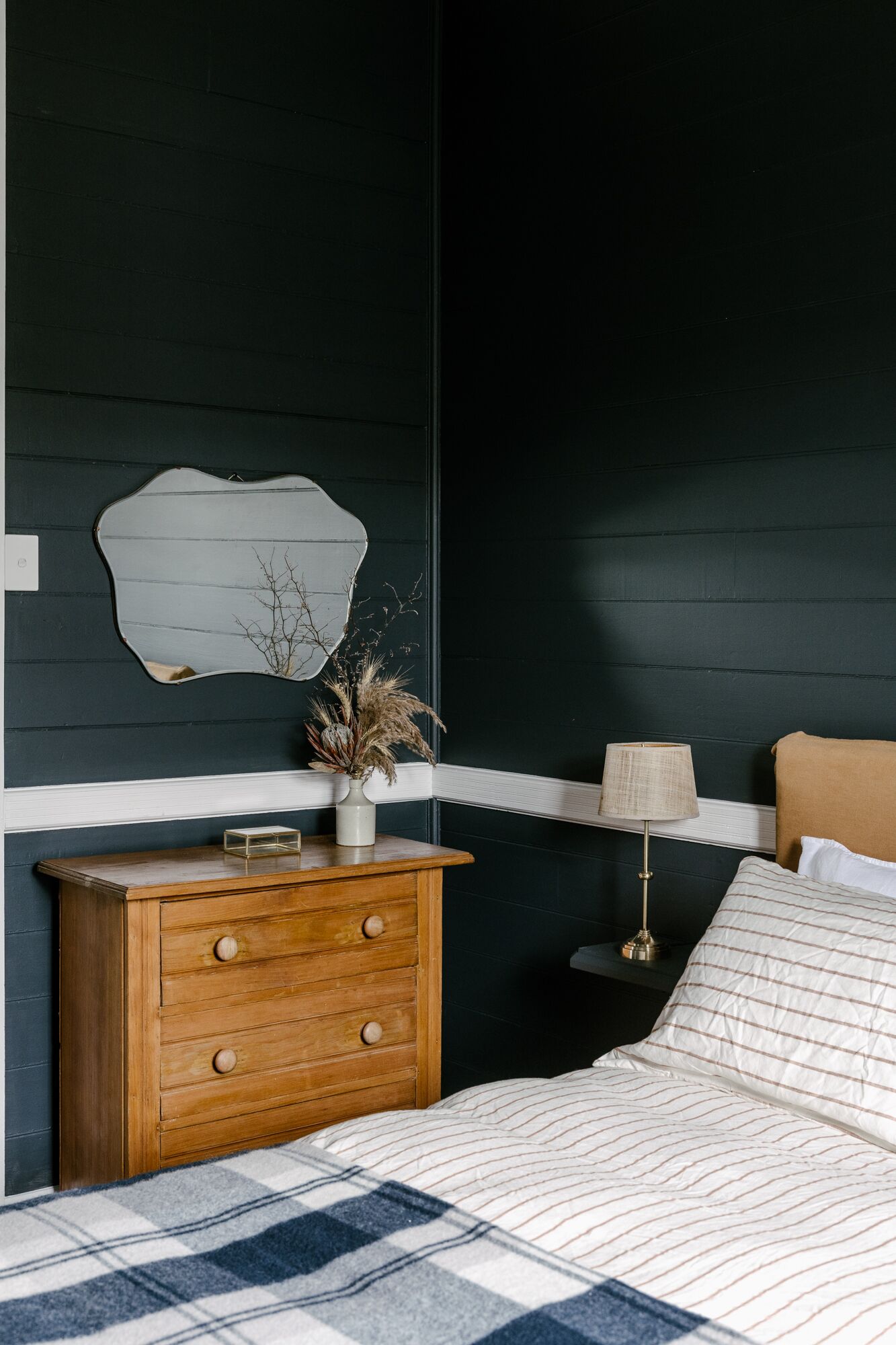Ein Schlafzimmer mit dunkler Wand, einer Holzkommode mit Schubladen, einem Bett mit gestreifter Bettwäsche, einer Lampe auf einem Nachttisch und einer Vase mit Trockenpflanzen auf der Kommode.