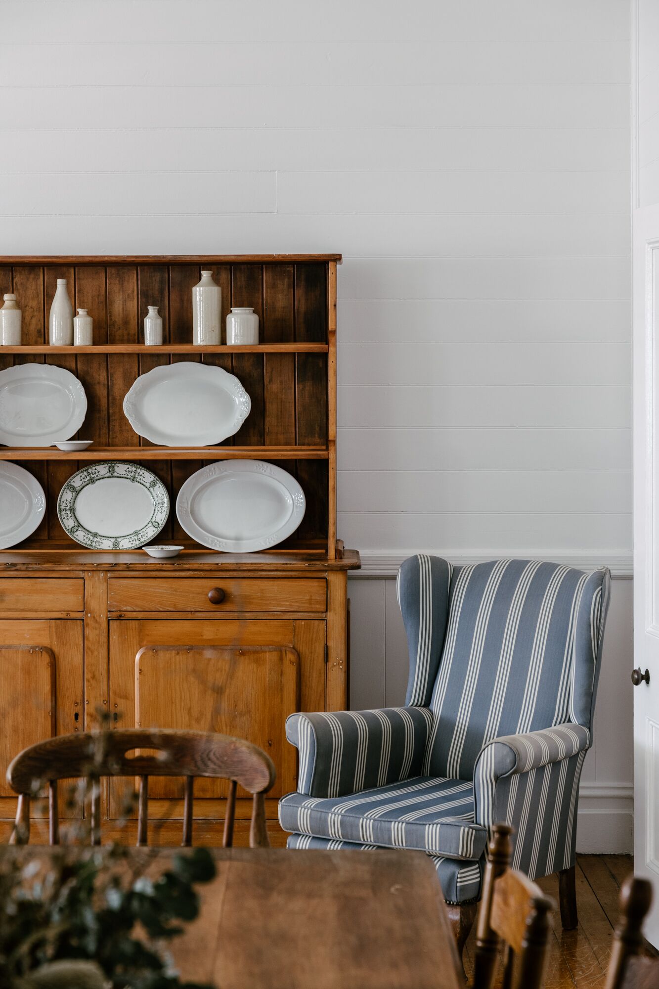 A dining area with a wooden cabinet displaying plates and jars, a striped armchair, and a wooden dining table.