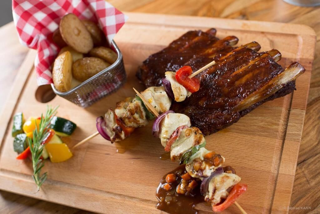 Une planche à découper en bois sur laquelle sont disposées des brochettes de poulet grillé, un bol de frites et un morceau de viande grillée.