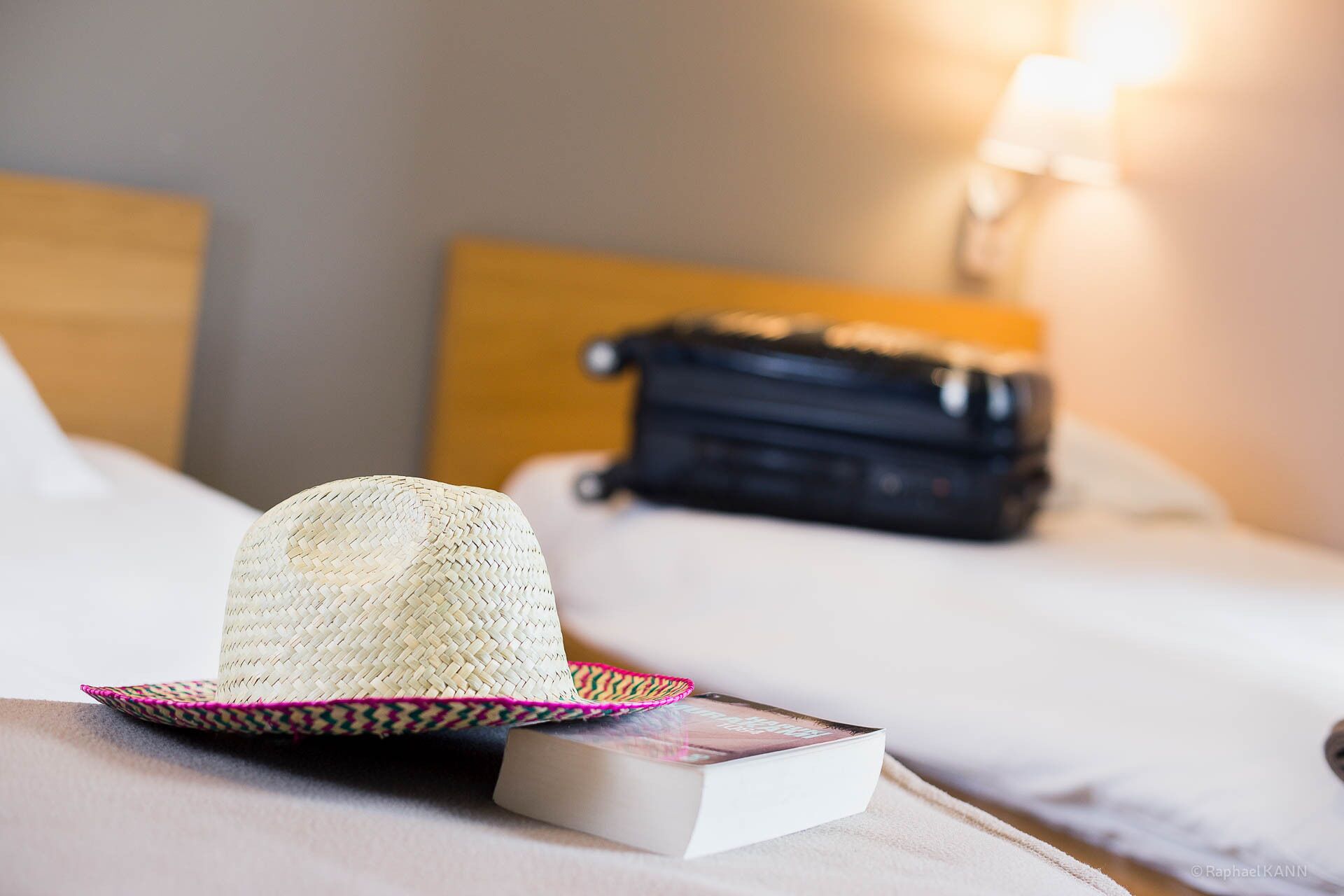 Un sombrero de paja sobre una cama, con un libro y un bolso negro al fondo.