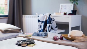 A neatly made bed with a folded towel, a book, a wallet, sunglasses, and a straw hat on a beige bedspread. A white desk with a plant and a framed picture is in the background. - Domaine de Garabaud (Mazeres)