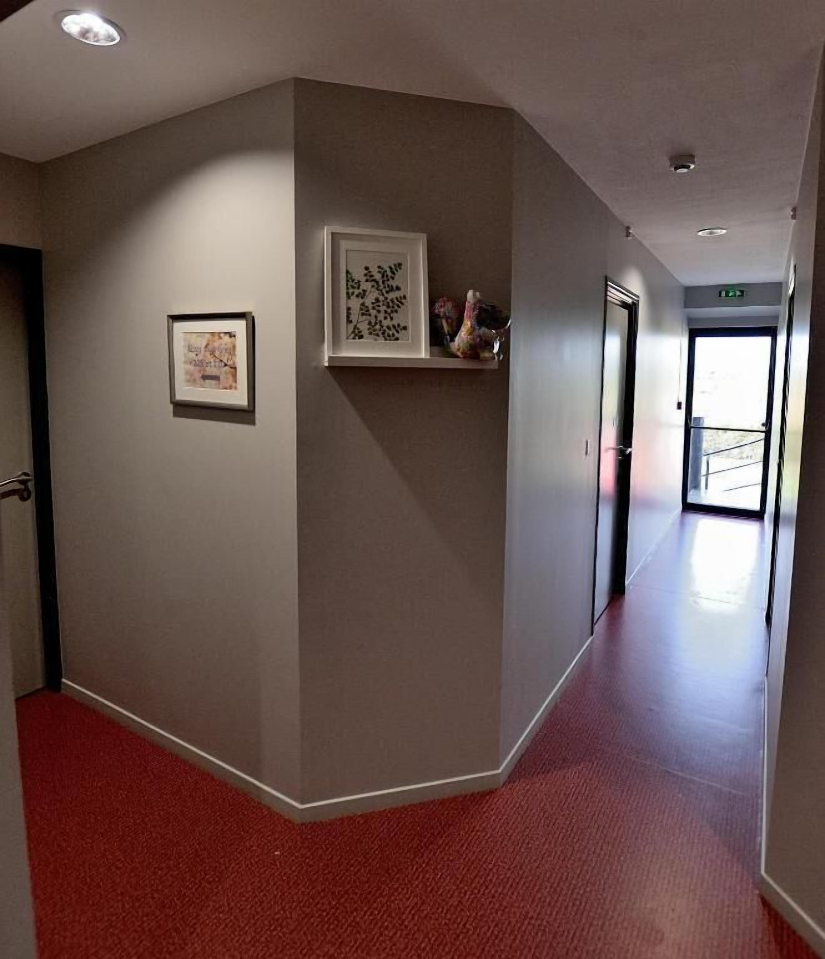 Um corredor com carpete vermelho, um quadro na parede e uma pequena estante com objetos decorativos.
