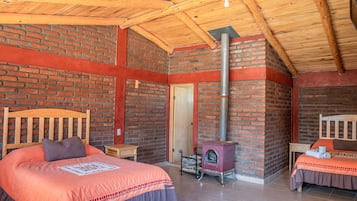 Quarto aconchegante com parede de tijolos, teto de madeira, cama com cabeceira, mesa pequena e lareira.