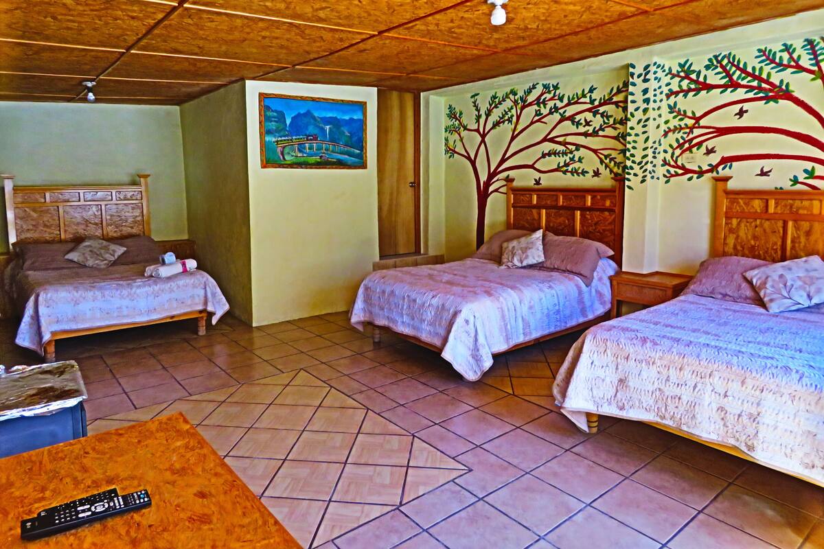 A hotel room with three beds, a wooden ceiling, a painting on the wall, and a tree mural.